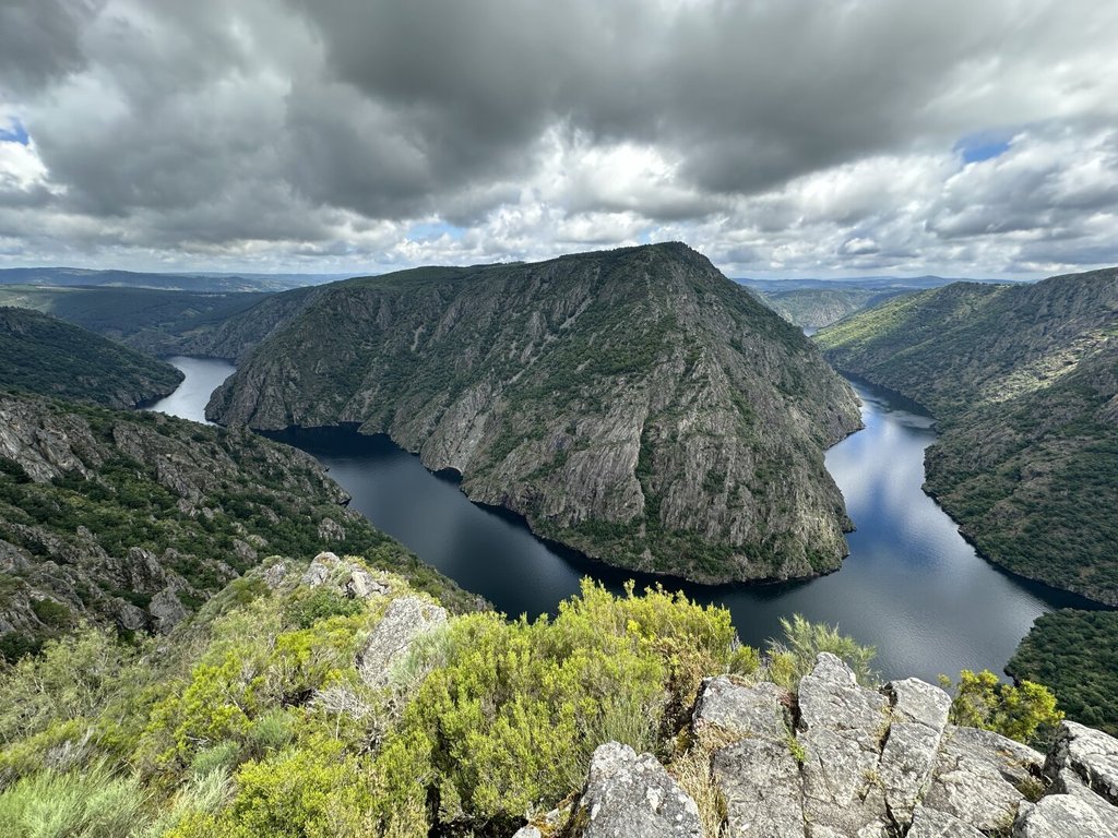Un meandro fluvial cerrado abraza una mole de monte cubierta de bosque denso; el cielo cargado se cierra sobre los riscos. En primer plano, la roca y el matorral asoman al borde del precipicio. La curva del agua precede a quien la mira por miles de años.