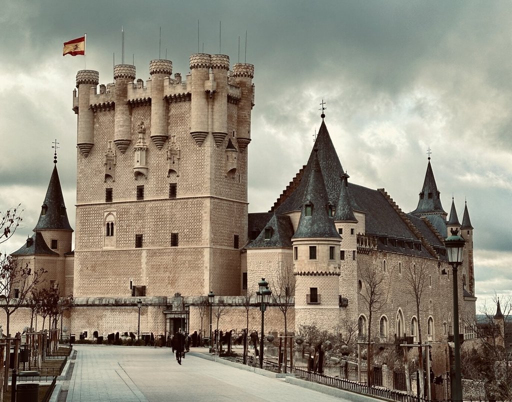 Alcázar fortificado sobre un espolón, torre del homenaje rematada en cubo con torrecillas almenadas y bandera en lo alto, cuerpo principal con tejados cónicos de pizarra a la derecha. Cielo plomizo, fila de árboles esqueléticos en el paseo. Más castillo de cuento que castillo de batalla: la silueta gana siempre a la función.