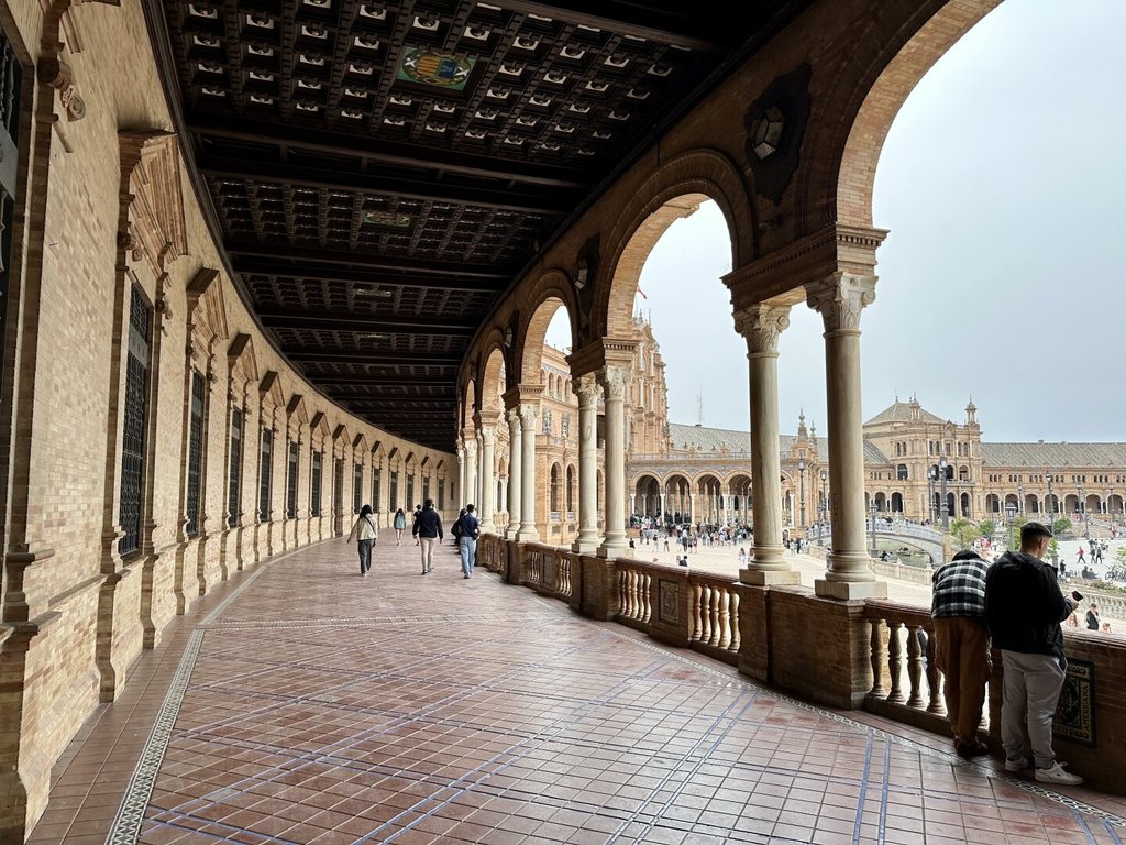Galería interior abovedada con techo artesonado polícromo y arcos de medio punto sobre columnas pareadas, suelo de baldosa rosa. Al fondo, gente paseando bajo la curva infinita; a la derecha, la fachada de la plaza con sus torres y arquerías. Arquitectura ceremonial pensada para impresionar a los suyos y a los de fuera: a un siglo de distancia, la cámara sigue cayendo en la trampa.