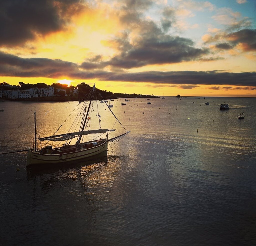 Velero tradicional amarillo amarrado en una bahía al amanecer, mástil sin velas izadas. Otros barcos puntean el agua tranquila hasta una costa lejana de casas blancas. El sol asoma entre nubes, derramando una franja dorada sobre el mar. Hay puertos donde el día empieza despacio porque el oficio sigue siendo esperar.