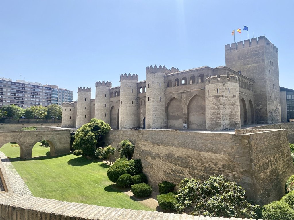 Palacio fortificado de muros amarillos con torres semicirculares almenadas y arquería ciega, una torre cuadrada más alta a la derecha con tres banderas en lo alto. Foso seco convertido en jardín, arcos de medio punto bajo el muro principal. Lo que fue residencia palaciega, prisión y cuartel hoy alberga otro tipo de poder: la piedra lleva mejor la rotación de funciones que sus inquilinos.