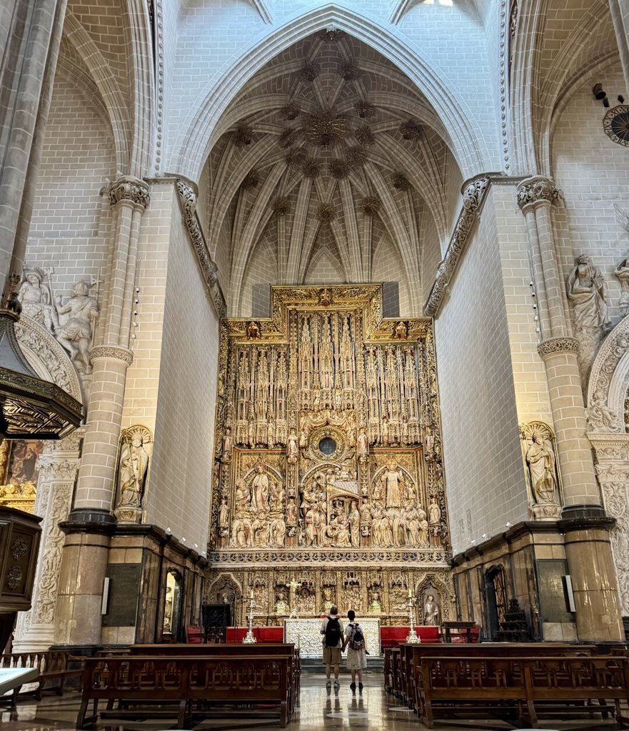 Bóvedas estrelladas sobre pilares cilíndricos y un retablo mayor que arde en oro al fondo, dos personas detenidas a su pie devueltas a su escala. Capillas laterales se asoman entre las columnas. La piedra blanca aspira hacia arriba, el oro tira hacia el mito.