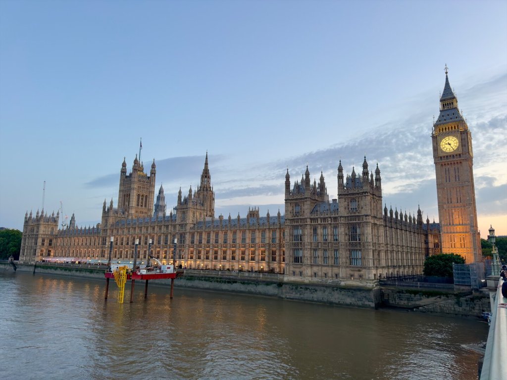 El palacio de Westminster visto desde el río al atardecer: una hilera larga de pináculos góticos color miel, ventanas ojivales y, a la derecha, la torre de Big Ben recibiendo todavía la última luz dorada cuando la fachada ya está en penumbra. El Támesis turbio en primer plano. El imperio sigue iluminado por hábito, no por necesidad.