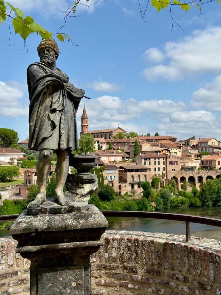 Estatua de bronce ennegrecido sobre un pedestal de piedra: un explorador con turbante, capa larga y bastón, mirando a un horizonte que ya no le incumbe. Detrás, un pueblo de tejados rojos baja hacia el río, con un campanario destacado y un puente de arcos. El monumento envejecido acaba siendo más parte del paisaje que figura sobre él.