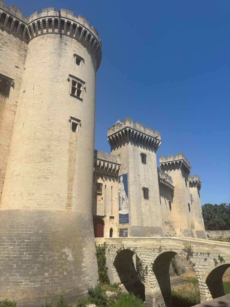 Castillo de piedra clara visto desde abajo: una torre redonda enorme a la izquierda y, detrás, varias torres cuadradas almenadas con saeteras estrechas como ojos pequeños. Un puente de piedra de tres arcos cruza un foso seco hasta la entrada. Cielo azul rotundo. Las fortalezas intactas son siempre una imagen de fracaso útil: las que no llegaron a tener que defenderse.