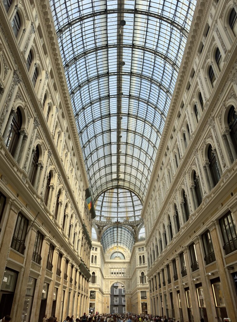 Interior de la Galleria Umberto I: bóveda inmensa de vidrio y hierro forjado dividida en cuadrículas regulares, dos plantas de fachadas eclécticas con balconadas y, en el fondo, una cúpula central que termina la perspectiva. Mucha gente diminuta abajo. El siglo XIX inventando catedrales sin santo, solo para que el comercio tuviera nave.