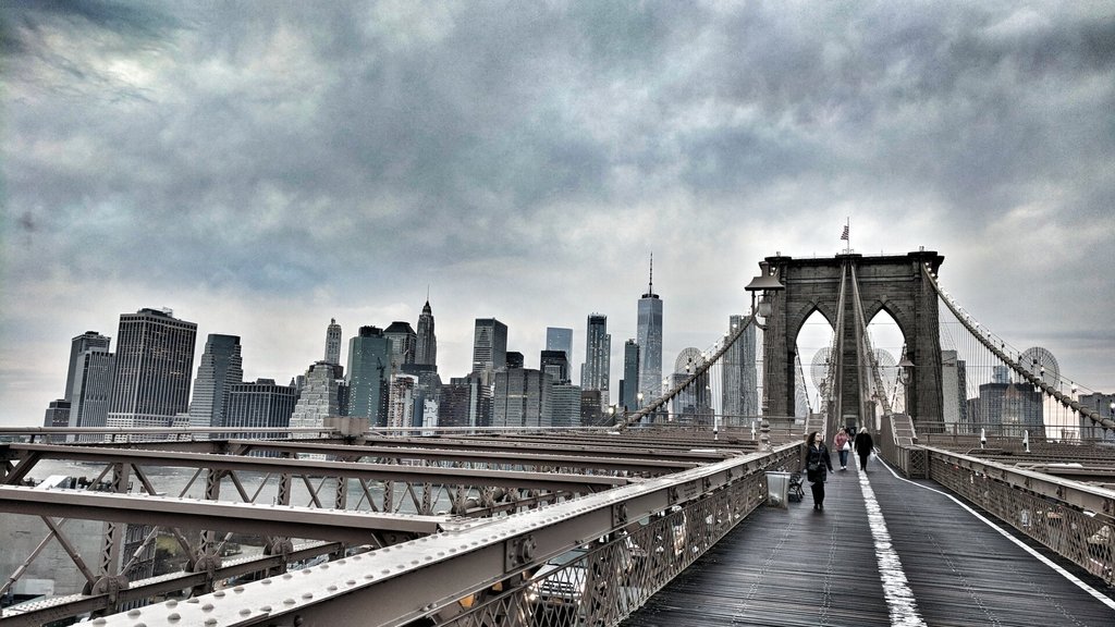 Pasarela peatonal del Puente de Brooklyn vista al frente: tablones gastados, cables verticales tensos hacia las almenas de piedra de la torre, una bandera diminuta en lo alto. Más allá, el skyline bajo un cielo de nubes pesadas y, a un lado, dos siluetas caminando hacia el otro lado. Cruzar es la forma más honesta de leer una ciudad.