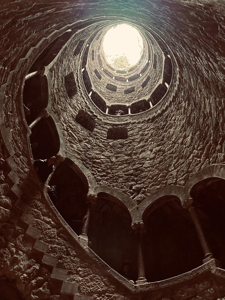 Pozo iniciático visto desde abajo: una espiral descendente de piedra labrada que sube en torno a un eje vacío hasta abrirse en un óculo de luz, con un árbol asomado al borde como advirtiendo que arriba aún hay mundo. En las galerías laterales se intuye gente diminuta. Bajar para ascender es solo gramática del mismo gesto.