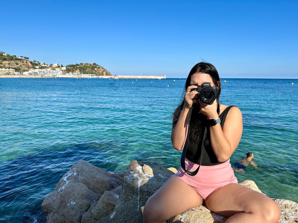 Mujer en bañador rosa sentada sobre una roca al borde del mar, descalza, levantando una cámara Sony para fotografiar al fotógrafo. Pelo al viento, agua color esmeralda detrás, una cabeza nadando a la derecha y una costa baja con caserío blanco al fondo. El visor le devuelve a quien la mira la mira de vuelta. Cuando la mirada se duplica, deja de haber sujeto y objeto.