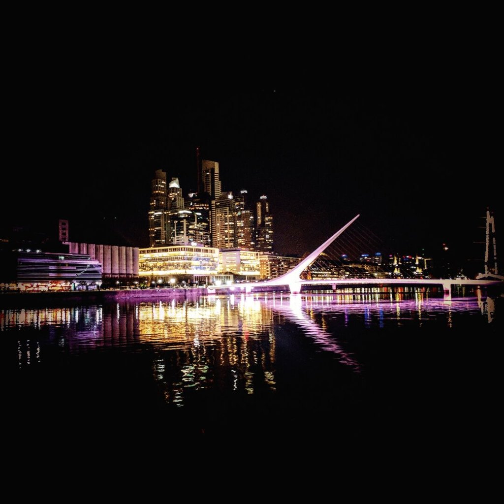 Skyline nocturno al otro lado de un río oscuro: torres de oficinas encendidas, una pasarela de mástil inclinado y tirantes blancos cruzando hacia ellas. El agua, casi negra, devuelve toda la ciudad invertida en hilos verticales de luz. La orilla se abstiene; deja que el reflejo haga el discurso. A veces una ciudad se entiende mejor por su réplica que por su original.
