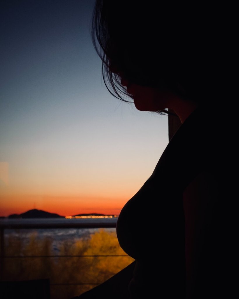 Silueta femenina recortada contra un ventanal: pelo, perfil bajado, hombro y curva del torso en sombra plena. Detrás, el horizonte se va degradando del naranja brasa al azul de noche, con una isla mínima y un puñado de luces lejanas haciendo collar sobre el agua. La oscuridad de dentro es más densa que la de fuera. Hay momentos en los que el cuerpo se vuelve frontera, no figura.