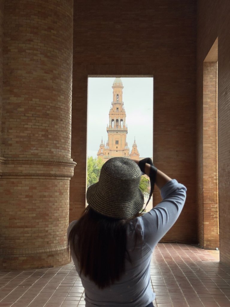 De espaldas, una mujer con sombrero de paja de ala ancha y melena larga fotografía un torreón de ladrillo visto a través de un vano arquitectónico. Las dos columnas macizas la enmarcan como visor de la propia escena. Toda foto incluye al fotógrafo aunque pretenda lo contrario.