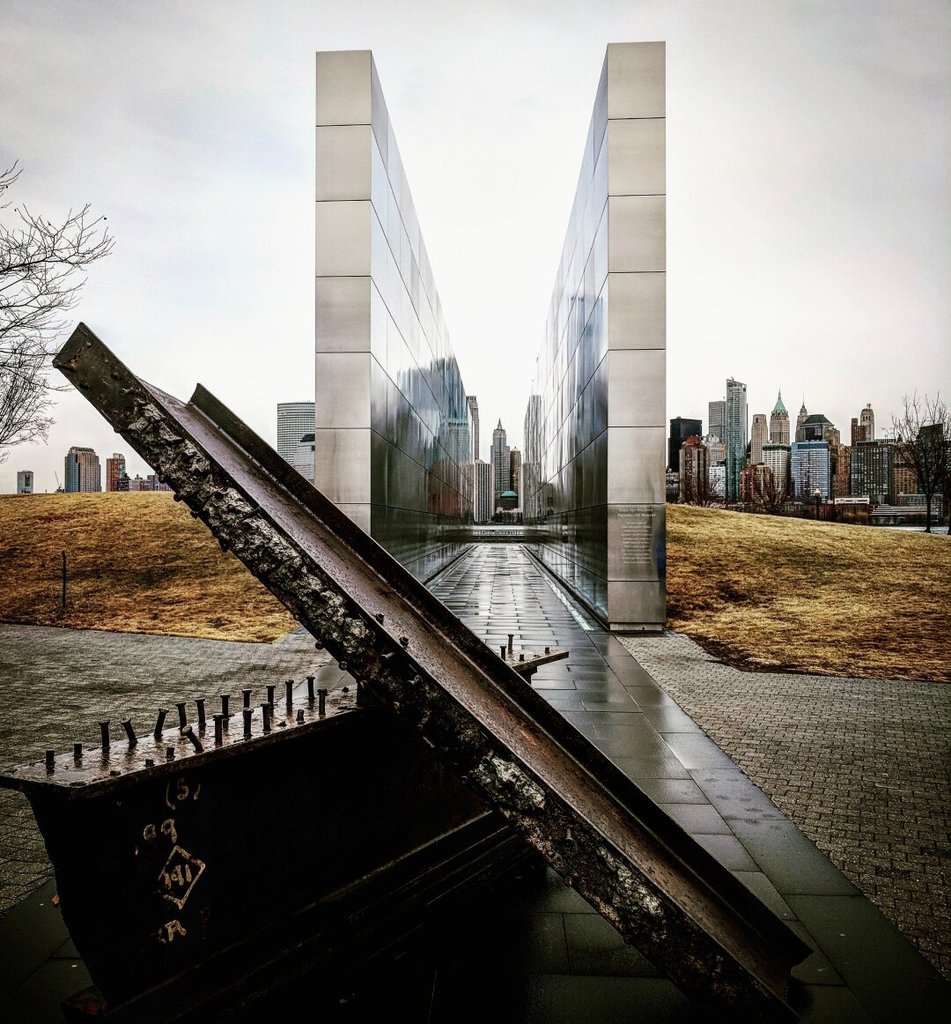 Monumento minimalista de dos muros de acero pulido entre los que se recorta una ciudad de rascacielos al fondo. En primer plano, una viga retorcida del derrumbe original, oxidada y conservada. La memoria también pide arquitectura.