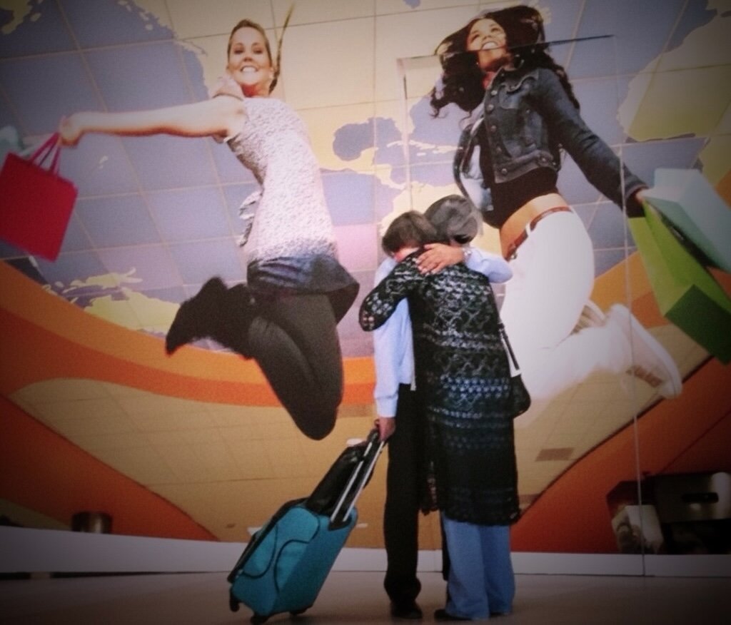 Aeropuerto: en primer plano, dos personas se abrazan junto a una maleta turquesa; detrás, un mural publicitario gigante con dos mujeres saltando con bolsas de compras sobre un mapa del mundo. El gesto verdadero queda más pequeño que el gesto inventado. Las despedidas reales nunca caben en un cartel.