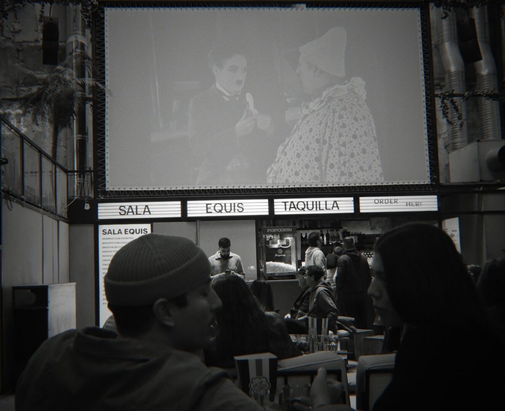 Interior de un cine-bar en blanco y negro: en lo alto, una proyección con Chaplin junto a un personaje encapotado en una escena del Klondike. Abajo, un rótulo retroiluminado anuncia SALA EQUIS, TAQUILLA, ORDER HERE, y dos cabezas en sombra ocupan el primer plano. Algunos lugares se llenan de gente porque la pantalla todavía las acompaña.