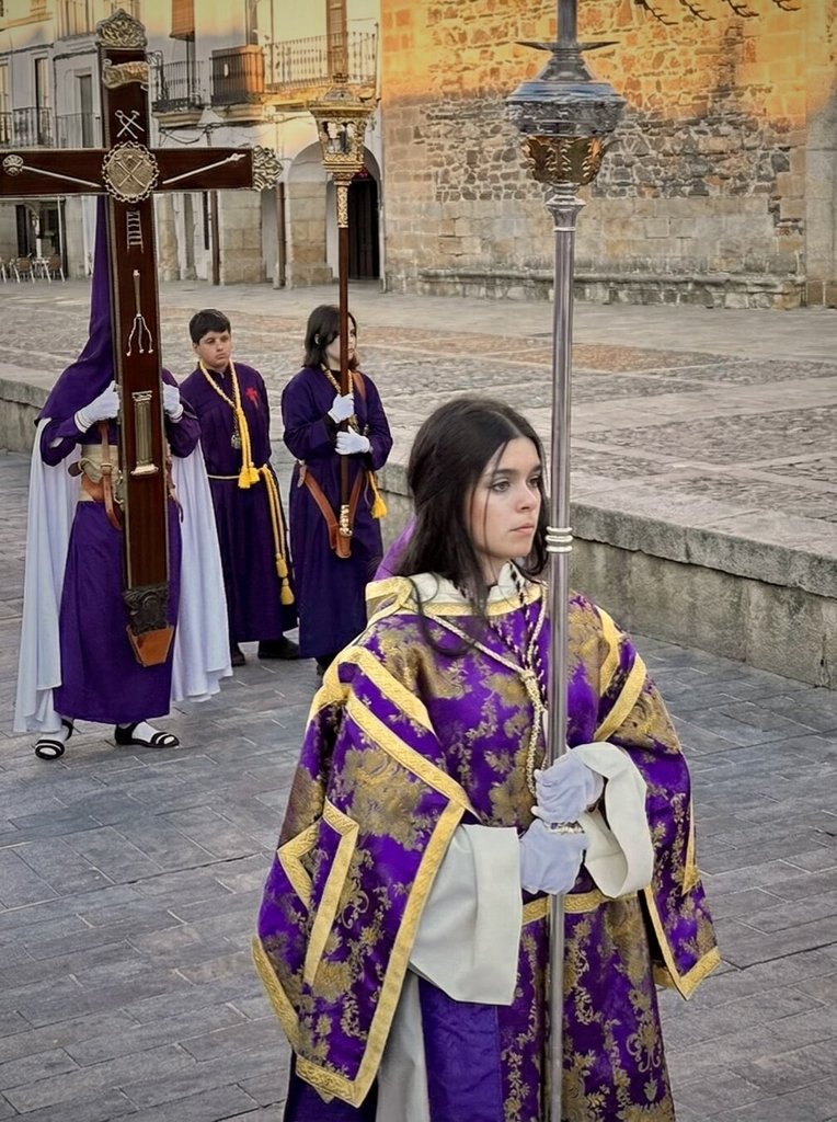 Cofradía en procesión al atardecer: en primer plano, una joven con dalmática púrpura bordada en oro sostiene un asta plateada con farol. Detrás, tres nazarenos en hábito morado portan la cruz, uno con capirote blanco. La piedra de la plaza recoge el oro de la última hora.