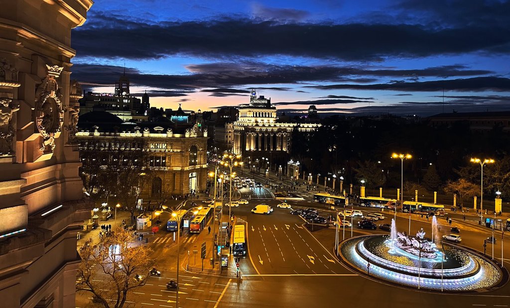 Plaza monumental al atardecer vista desde un balcón en altura, con fuente iluminada y una avenida de tráfico rodeándola. Las cúpulas y palacios al fondo recortan el último naranja del horizonte. Sólo la hora azul vuelve elegante hasta el atasco.