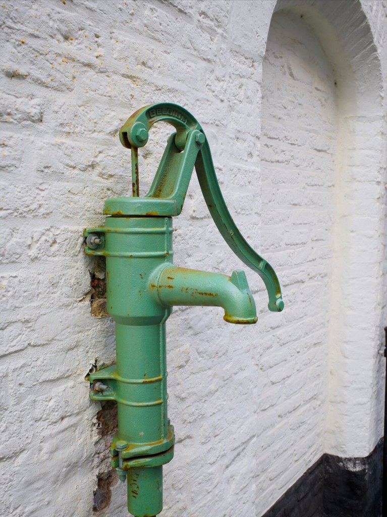 Bomba de agua manual antigua, pintada en verde mar y oxidada en las juntas, sujeta a una pared encalada con tornillos pasantes. La palanca curva descansa hacia abajo y un caño grueso espera el siguiente cubo. La pieza sigue ahí décadas después de que el grifo de la cocina haga el trabajo.