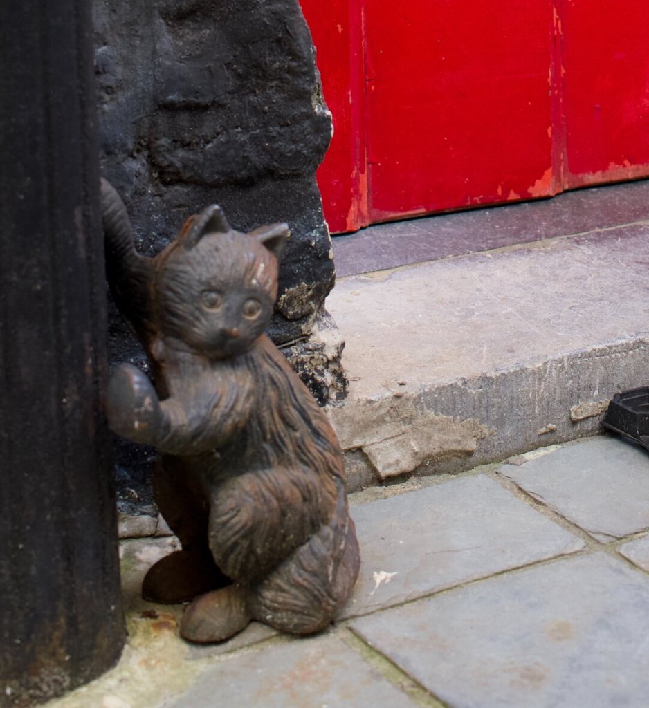 Un raspador de barro en forma de gato pequeño, fundido en hierro oscuro y oxidado, se apoya contra el quicio de piedra negra junto a un poste también de hierro. Detrás se asoma el panel rojo brillante de una puerta. El gato lleva décadas haciéndose el sorprendido sin que nadie le pase la mano por encima.