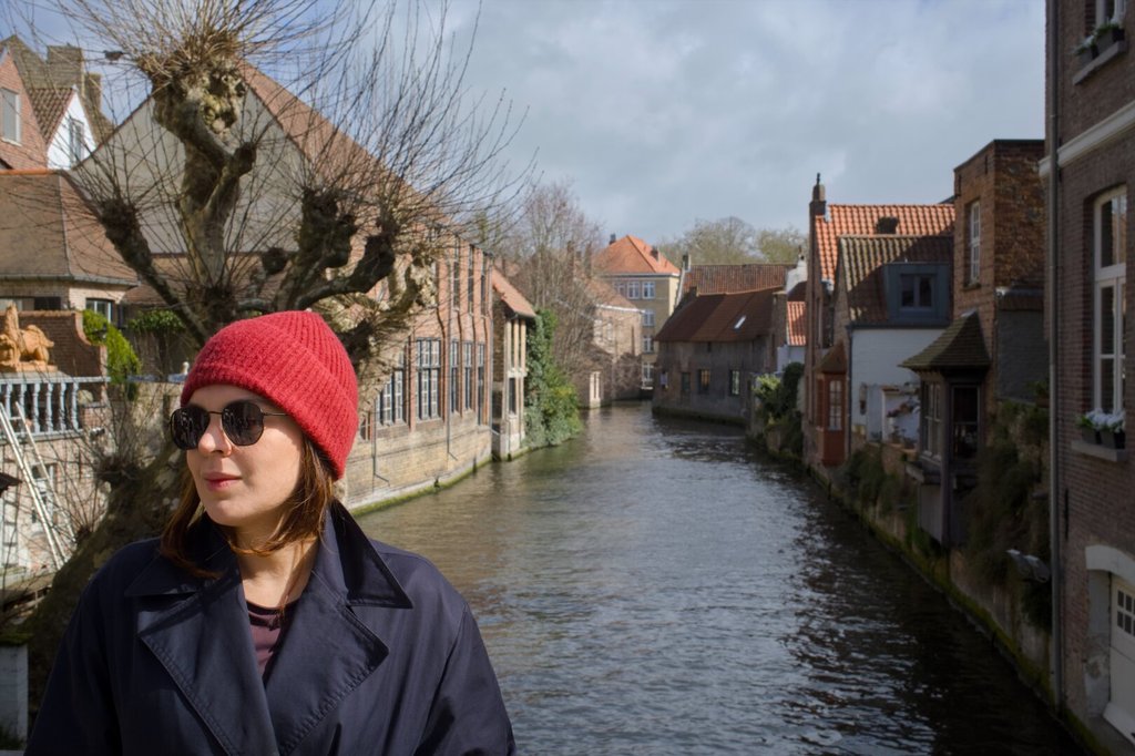 Una mujer joven con gorro rojo de lana y gafas de sol mira hacia el lado, en perfil de tres cuartos; lleva una gabardina azul oscuro abierta sobre una camiseta morada. Detrás, un canal estrecho ordena la perspectiva entre casas de tejado a dos aguas y árboles podados en bola. El gorro concentra todo el calor del cuadro.