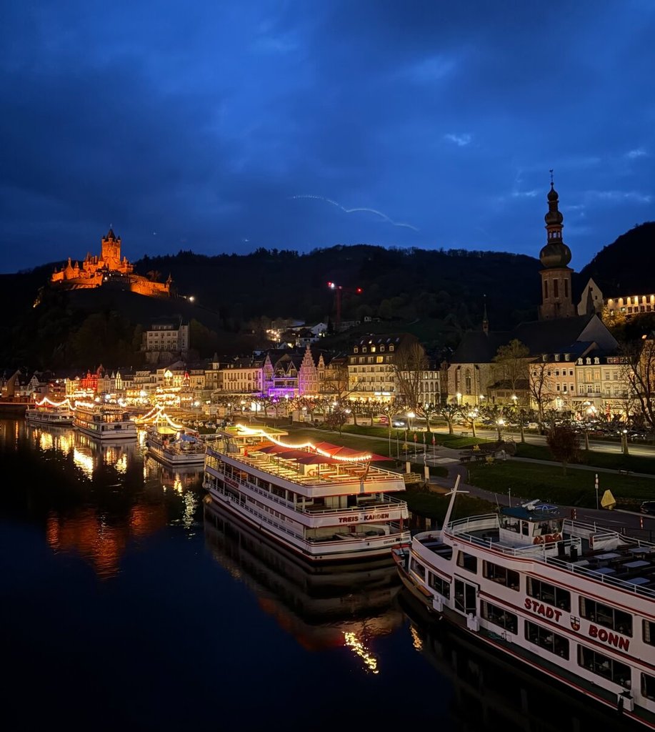 Pueblo a orillas de un río al anochecer: en lo alto, un castillo iluminado de naranja preside la silueta del valle; abajo, dos barcos de pasaje amarrados se reflejan en el agua negra. Entre el castillo y el muelle, casas con luces interiores y una iglesia de campanario bulboso ordenan la línea del paseo. La hora azul aguanta mientras los barcos esperan a que alguien suba.