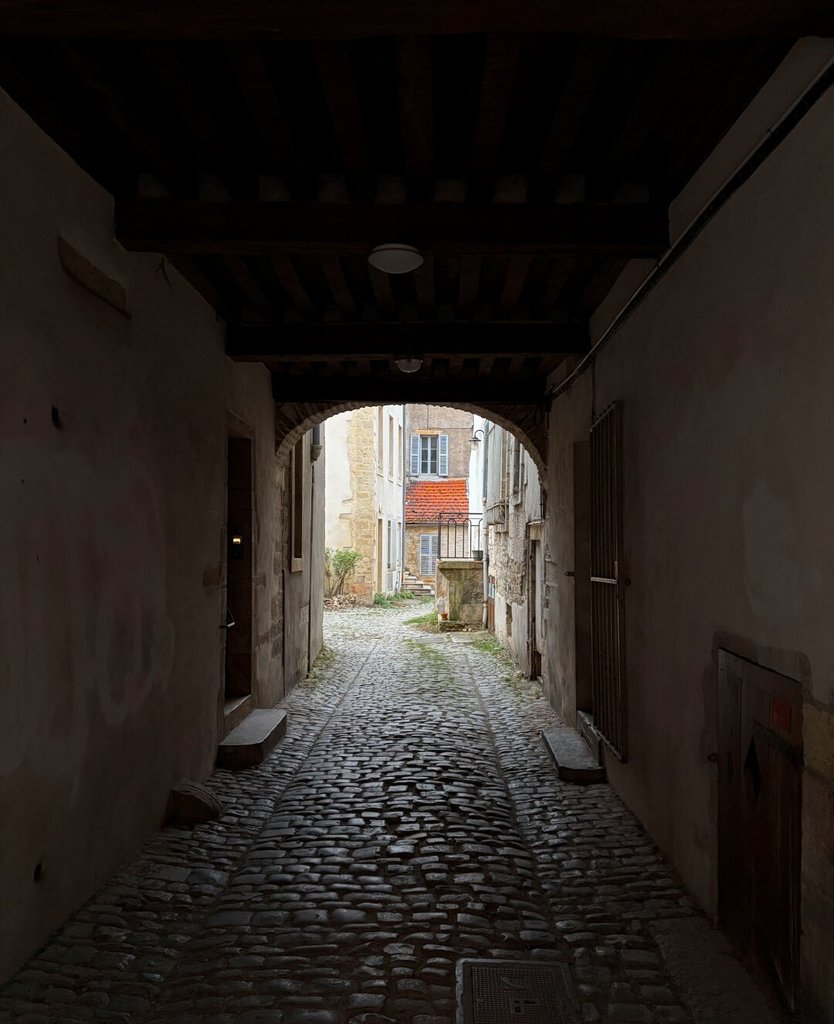 Pasaje cubierto con techo de vigas de madera oscura y paredes encaladas; el suelo de adoquines pulidos baja en pendiente suave hacia un patio iluminado al otro extremo. Al fondo, fachadas claras de tejado rojizo aparecen recortadas por el arco de salida. El umbral hace de visor: encuadra el día que viene desde fuera.