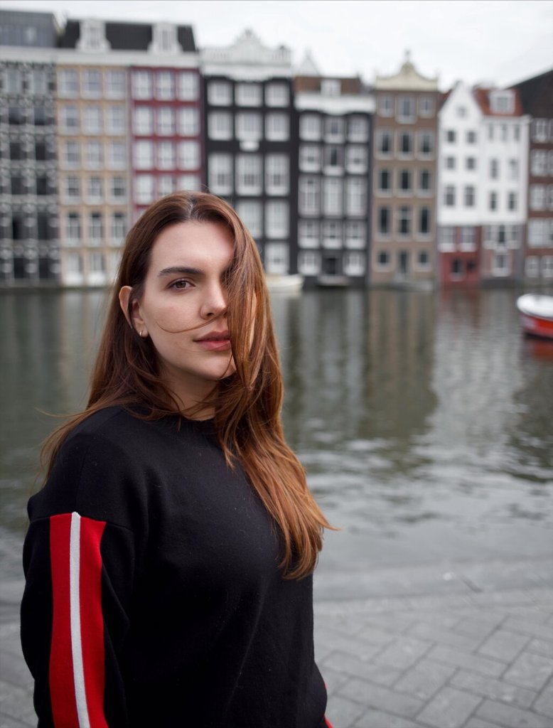 Retrato de una mujer joven; un mechón cruza su cara y los ojos se sostienen en cámara sin sonrisa de cortesía. El suéter negro tiene una franja roja vertical en el hombro. Detrás, fachadas estrechas y altas se alinean en hilera sobre un canal de aguas quietas, con un bote rojo asomando al margen.