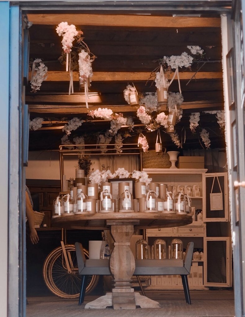 Desde la calle, una puerta abierta deja ver el interior de una tienda en Segovia: ramas de flores de cerezo cuelgan de las vigas de madera sobre una mesa redonda cargada de velas y frascos. A la izquierda, el brazo de una persona apoyada en una bicicleta antigua entra en el cuadro sin terminar de aparecer. El umbral ofrece la escena y a la vez la retiene tras el marco.