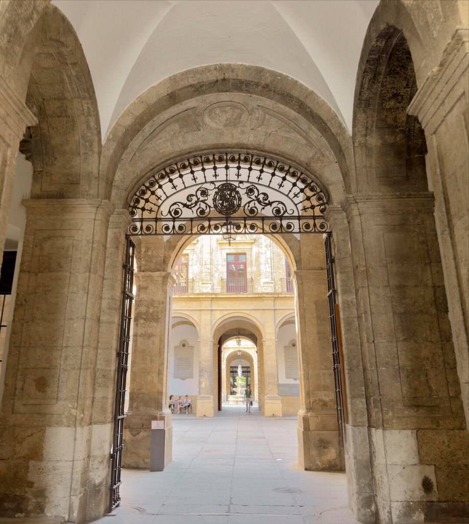 Vano de piedra en sombra enmarca una reja de forja con un escudo en el centro, abierta hacia un patio interior bañado de sol. Al fondo, arcos y un balcón ocre, y figuras menudas sentadas o de paso que dan la escala. El umbral en penumbra cede a la claridad del patio, como una bisagra entre dos densidades de luz.