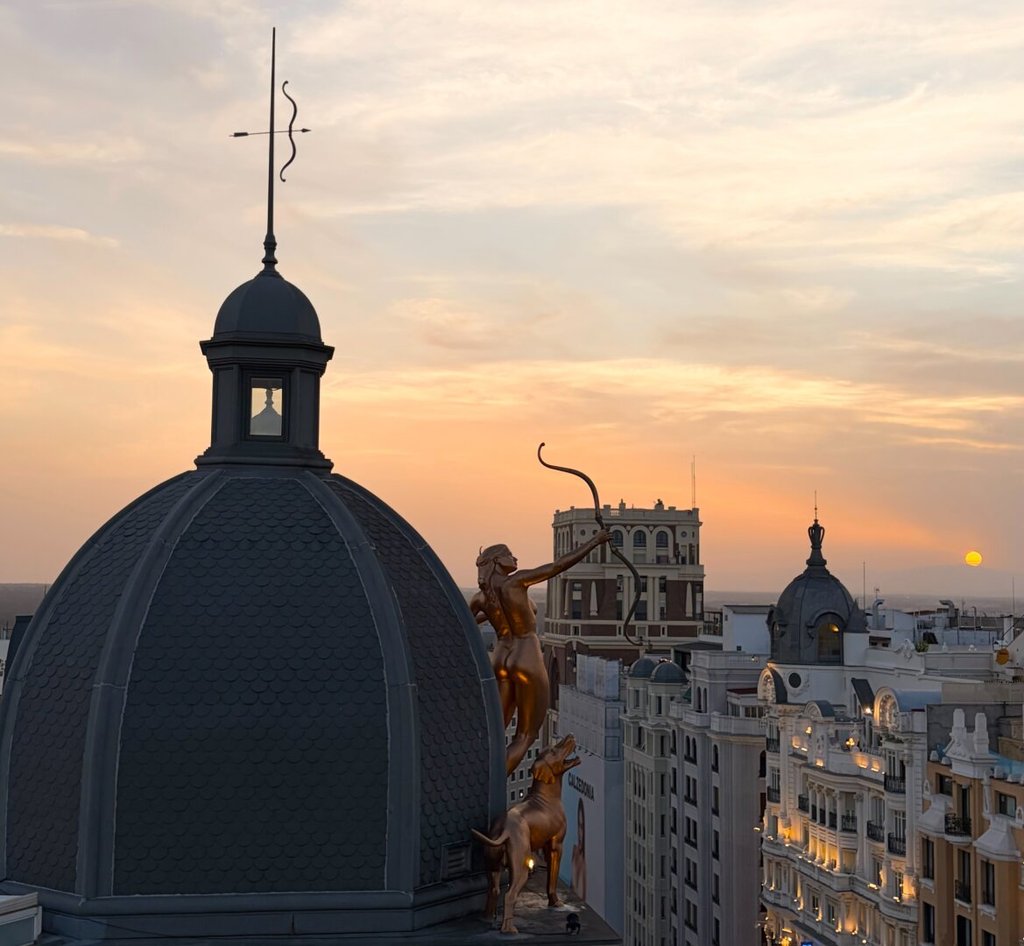 Sobre los tejados, una cúpula de pizarra oscura rematada por un pararrayos con forma de serpiente y flecha; al lado, una arquera dorada y desnuda tensa el arco hacia el cielo, con un perro a sus pies. Apunta al sol, que ya es un disco naranja a ras del horizonte y se pone solo, sin que nadie tenga que derribarlo. Abajo, las fachadas encienden las ventanas mientras el día cede.
