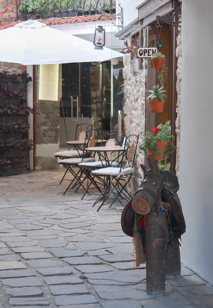 Terraza de un café en un callejón empedrado: parasol blanco, mesas de bistró con cojines, macetas de geranios y un cartel de OPEN junto a un farol viejo. En primer plano, un caballo hecho con un tronco lleva puesta una silla de montar de cuero y una brida de cuentas, ensillado y sin jinete. Un animal de mentira esperando un viaje que no toca.