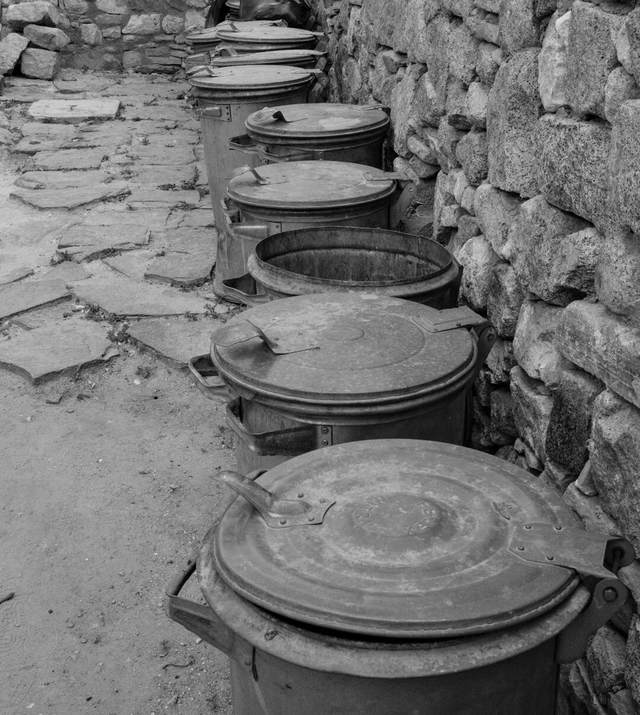 Una hilera de cubos de basura metálicos, abollados y con tapa, se alinea contra un muro de piedra a lo largo de un camino empedrado. Todos cerrados menos uno, destapado, que rompe la formación como quien se queda fuera de fila. El metal gastado se repite igual que un rosario de objetos que nadie mira y todos usan.