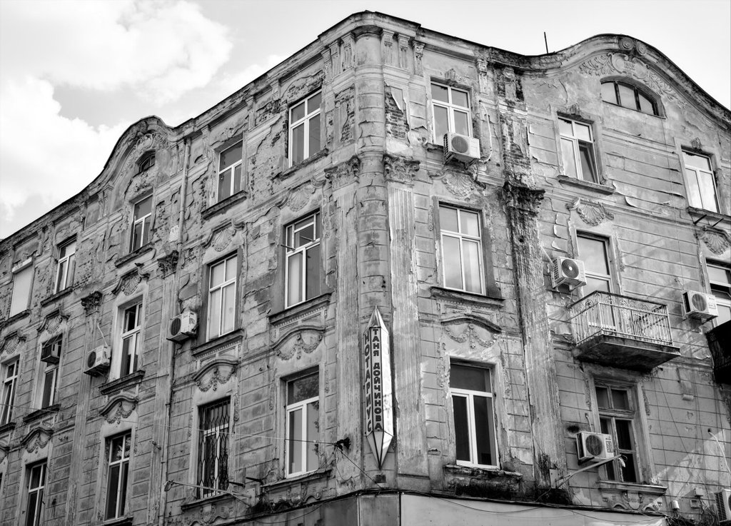 Edificio antiguo de esquina, de molduras y guirnaldas de yeso, con el revoque caído en placas que dejan ver el ladrillo. Sobre la fachada noble cuelgan aparatos de aire acondicionado y un rótulo vertical de notaría, y un balcón de hierro se asienta torcido. La grandeza de otro siglo cediendo, piso a piso, a los arreglos de este.
