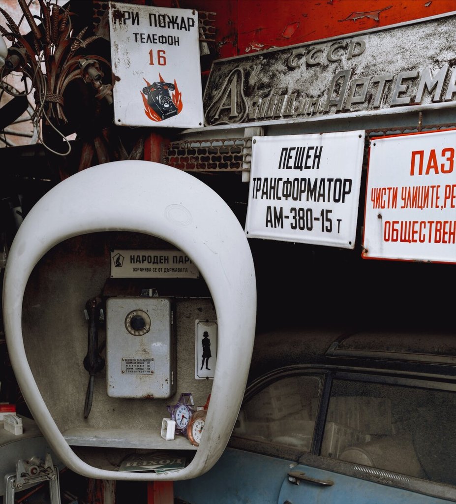 Una cabina telefónica blanca con forma de huevo guarda un teléfono de disco gris, montada sobre un mercadillo de chatarra de la era soviética. Encima se amontonan rótulos esmaltados en cirílico, un aviso de incendios con su teléfono en llamas, relojes parados y el morro de un coche viejo. Tanta máquina para hablar con alguien, toda muda y apilada contra la pared.