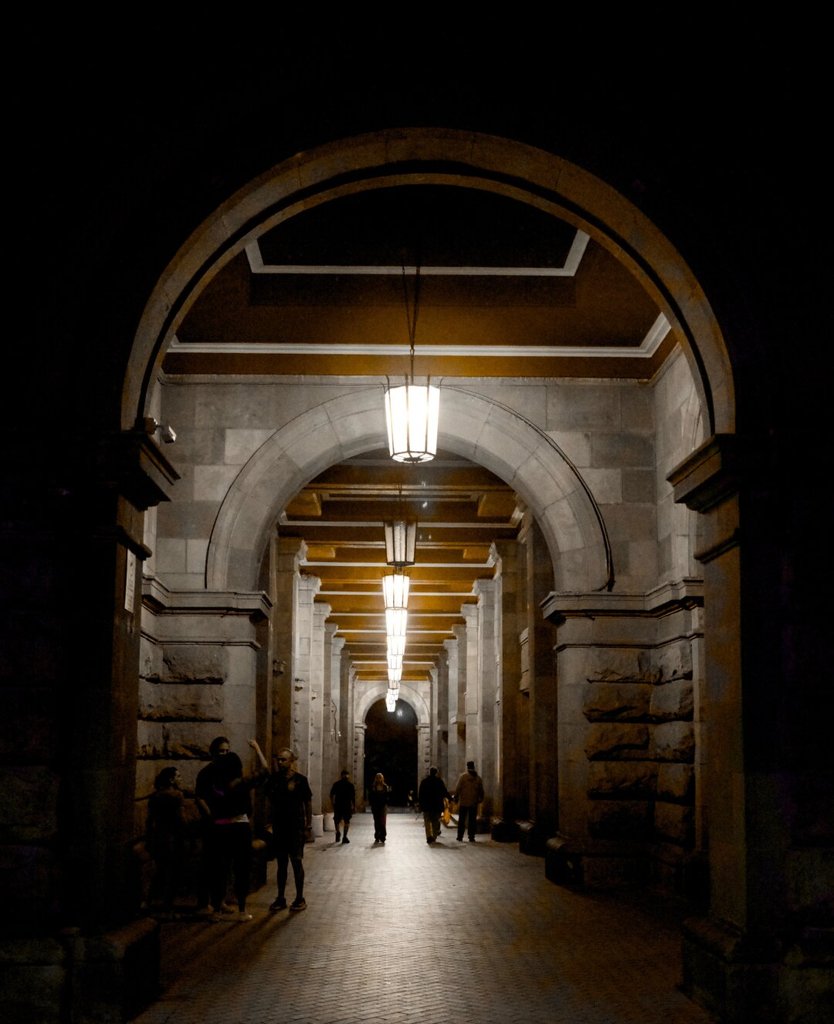 Un gran arco de piedra enmarca la boca de un pasaje abovedado, con un farol colgando en el centro y más faroles que se pierden en hilera hacia el fondo. Bajo la arcada, un grupo conversa a la izquierda mientras otras figuras caminan hacia el extremo oscuro, pequeñas entre los pilares. La gente entra por el umbral y el corredor se la va llevando.