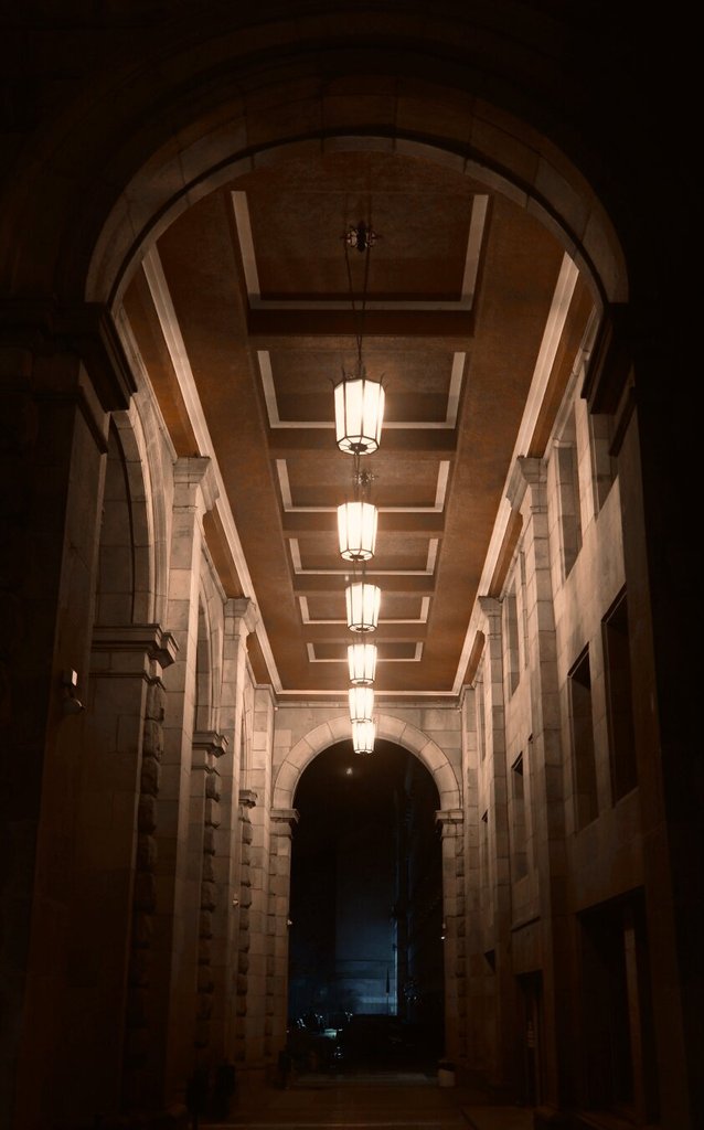 Un pasaje cubierto, de noche, alinea sus faroles colgantes bajo un techo de casetones que se estrecha en perspectiva. A los lados, pilastras de piedra almohadillada, pesadas y repetidas, sostienen la bóveda; al fondo, un arco se abre a una calle apenas iluminada donde se adivinan coches. La luz ordena el tránsito hacia lo que no se ve.