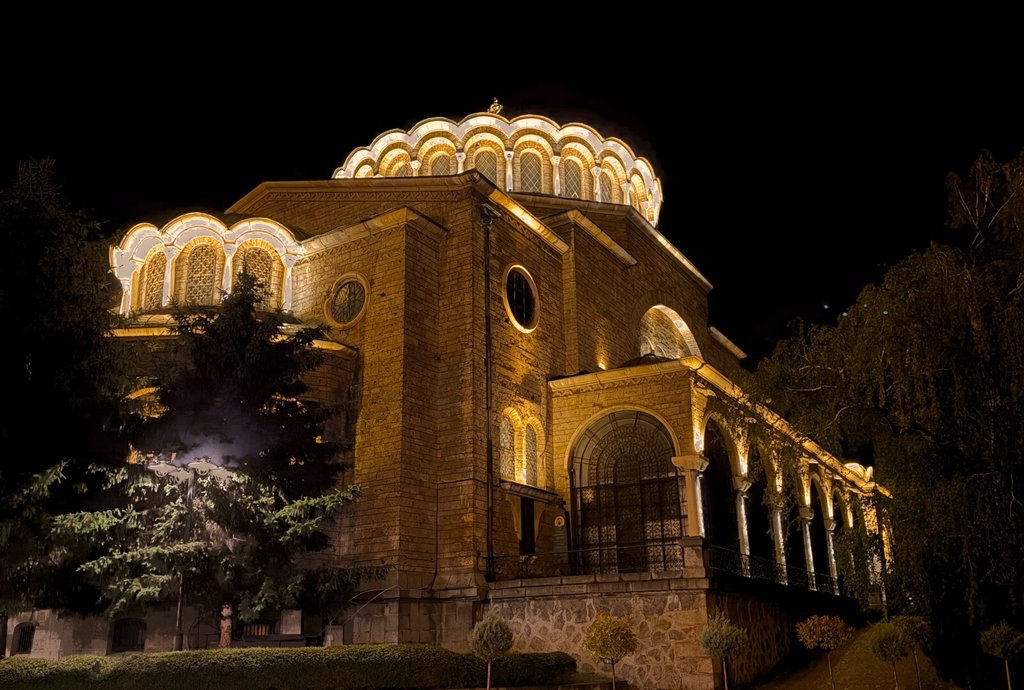 Una iglesia de piedra dorada se enciende sola contra un cielo completamente negro. La cúpula, festoneada y perfilada con hileras de bombillas, corona un cuerpo de ventanas en arco y un pórtico de columnas finas a la derecha; abajo, escalones y arbustos recortados suben hacia ella entre árboles oscuros. Alrededor, la noche se ha llevado todo lo demás.