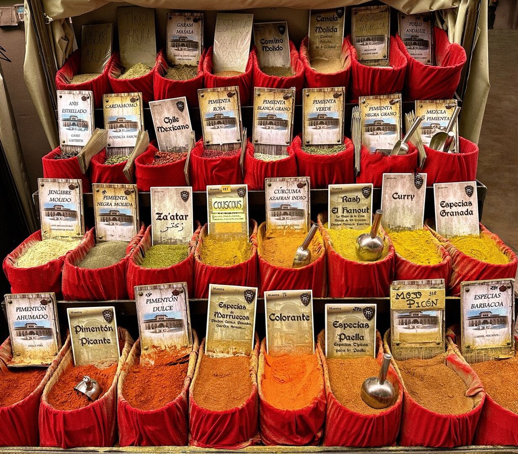 Un mostrador de especias se despliega como un pequeño imperio de polvo y color, con sacos, cajas y latas alineadas bajo una luz de mercado que no aclara del todo. Lo que fija la mirada no es el orden, sino la tensión entre lo que se vende y lo que se guarda, como si cada paquete tuviera un temblor de supervivencia.