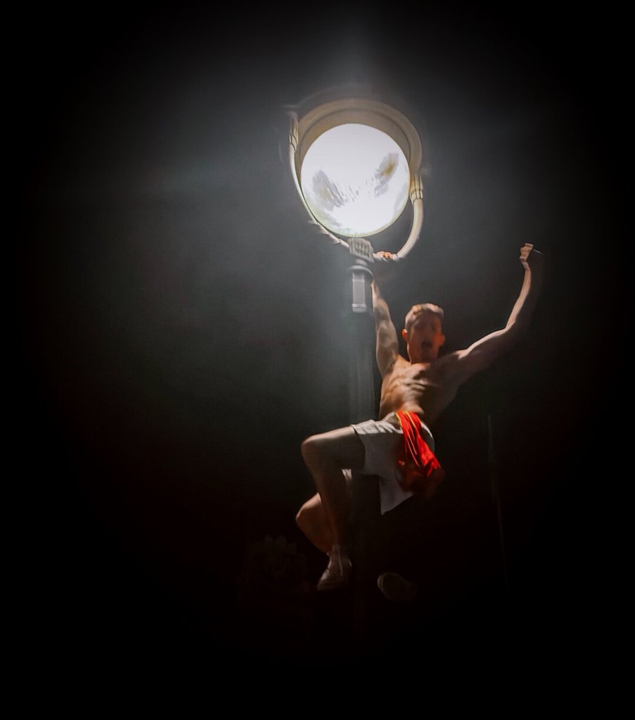 Un hombre sin camiseta se agarra a una farola de globo clásico en plena oscuridad; toda la luz sale del propio farol, que le talla el torso y deja el resto en negro. Una pierna encaja contra el fuste, un brazo se estira hacia arriba, una bandera de España cuelga de la cintura. El cuerpo queda suspendido entre trepar y caer, sostenido solo por el gesto.