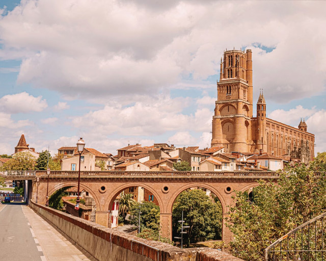 La catedral de ladrillo de Albi domina tejados bajos y un puente de arcos sobre árboles.