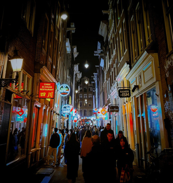Una calle estrecha de Amsterdam reúne peatones bajo farolas y rótulos de neón de restaurantes asiáticos.