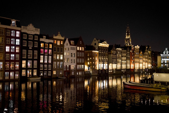 Casas estrechas iluminadas bordean un canal nocturno de Amsterdam, con reflejos dorados y una embarcación amarrada.