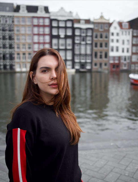 Una mujer de cabello largo mira a la cámara junto a un canal de Ámsterdam, con casas estrechas al fondo.