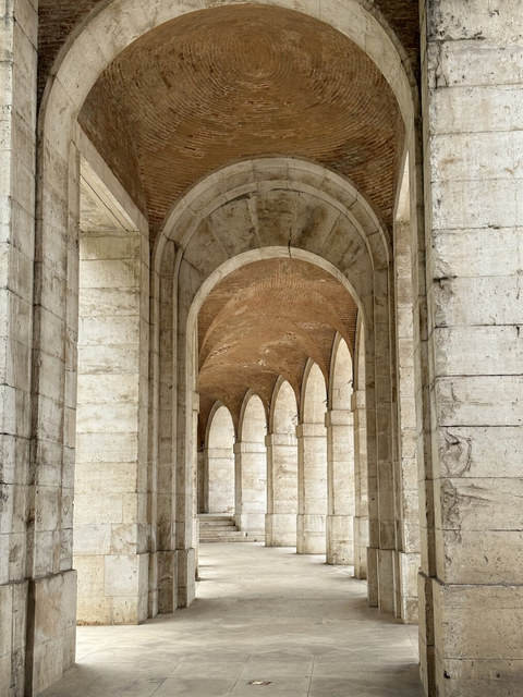 Arcos de piedra sucesivos enmarcan una galería abovedada de ladrillo con suelo claro y luz lateral.