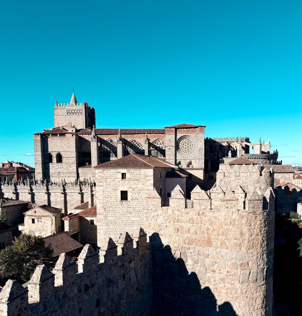 La catedral de Ávila sobresale tras las almenas de la muralla bajo un cielo azul despejado.