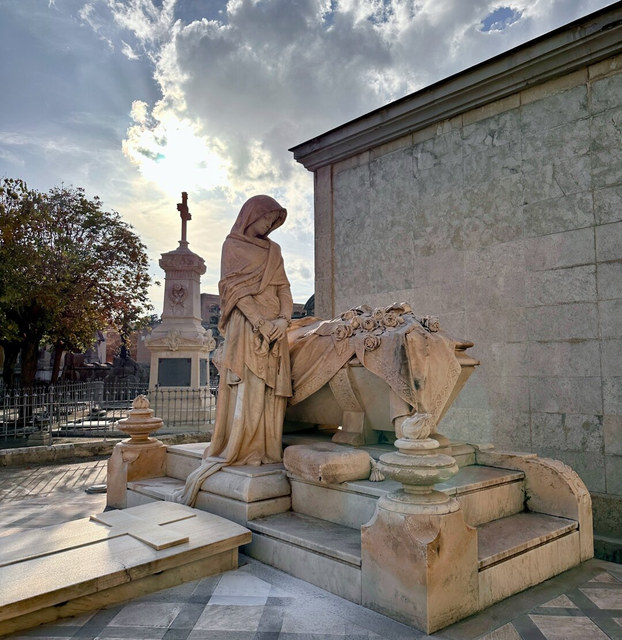 Una escultura funeraria representa a una mujer inclinada junto a una tumba en un cementerio de Barcelona.