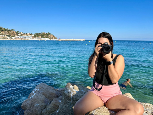 Una mujer sentada sobre rocas apunta su cámara hacia el objetivo, con un nadador detrás en el mar.