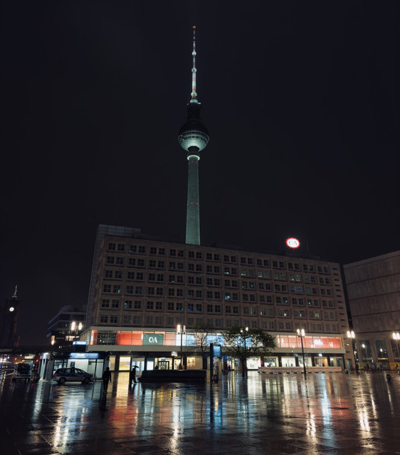 La torre de televisión de Berlín se eleva iluminada sobre una plaza mojada y un edificio comercial.