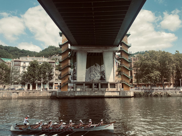 Una trainera con remeros avanza bajo un puente urbano sobre la ría de Bilbao.
