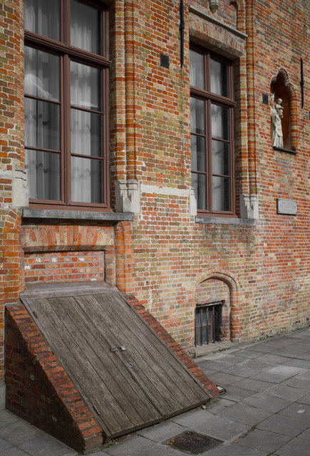 Una fachada de ladrillo en Brujas combina ventanales altos, una puerta inclinada de madera y una pequeña estatua en hornacina.